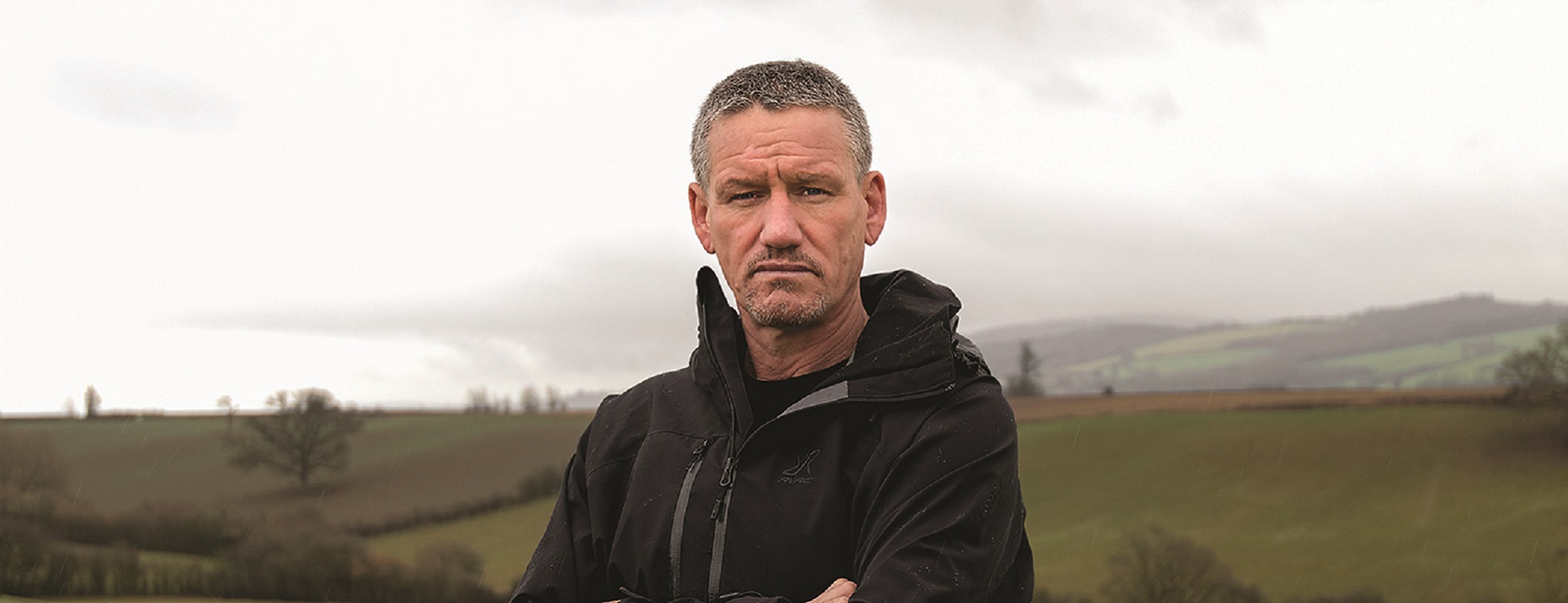 Billy Billingham standing with his arms folded in a field with hills in the background