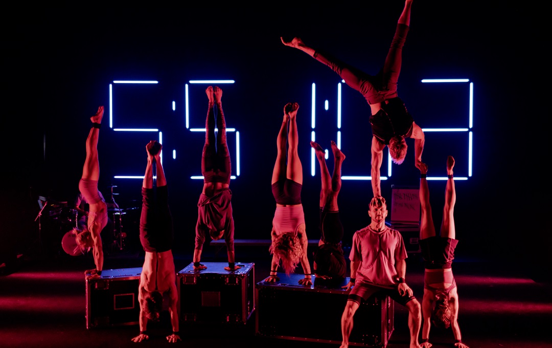 Acrobats performing on a dark stage in low red light with a digital time stamp behind them.