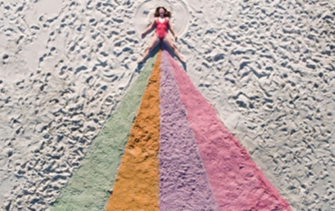An aerial photo of Lucy Peach lieing on a beach with multi-colour sand underneath her legs