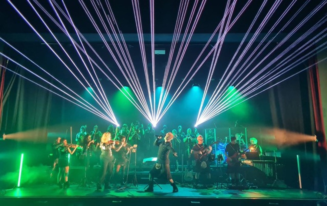 A band performing with an orchestra with white lasers overhead